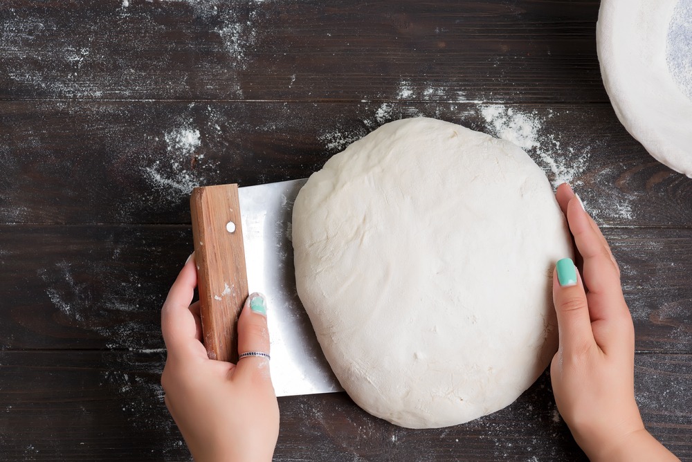 Hands shaping dough with a wooden-handled dough scraper on a floured surface.